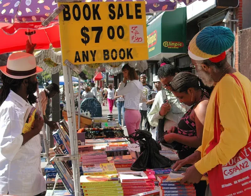 People browse books at an outdoor Harlem book fair table beneath a hand-painted book sale sign.