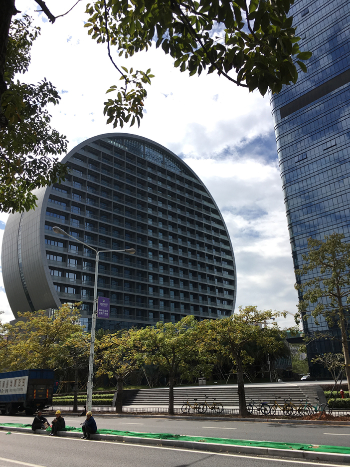 A tall building with a rounded, arch-like outer profile stands beside a glass tower outside the Sino-Finnish Design Park. Trees and parked bicycles line the street below.