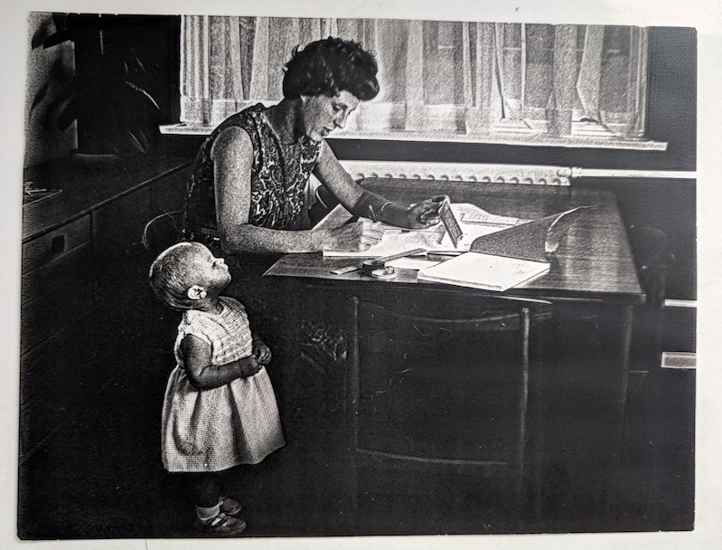Ann Moffatt writes on papers spread across her kitchen table while her young child stands beside her and looks up at her.