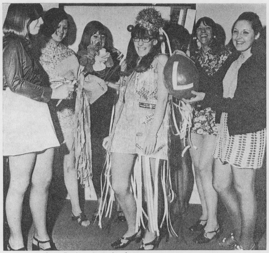 Anne Davis stands among smiling coworkers at her leaving party, wearing a dress made from long strips of punched paper tape and holding a balloon marked L.