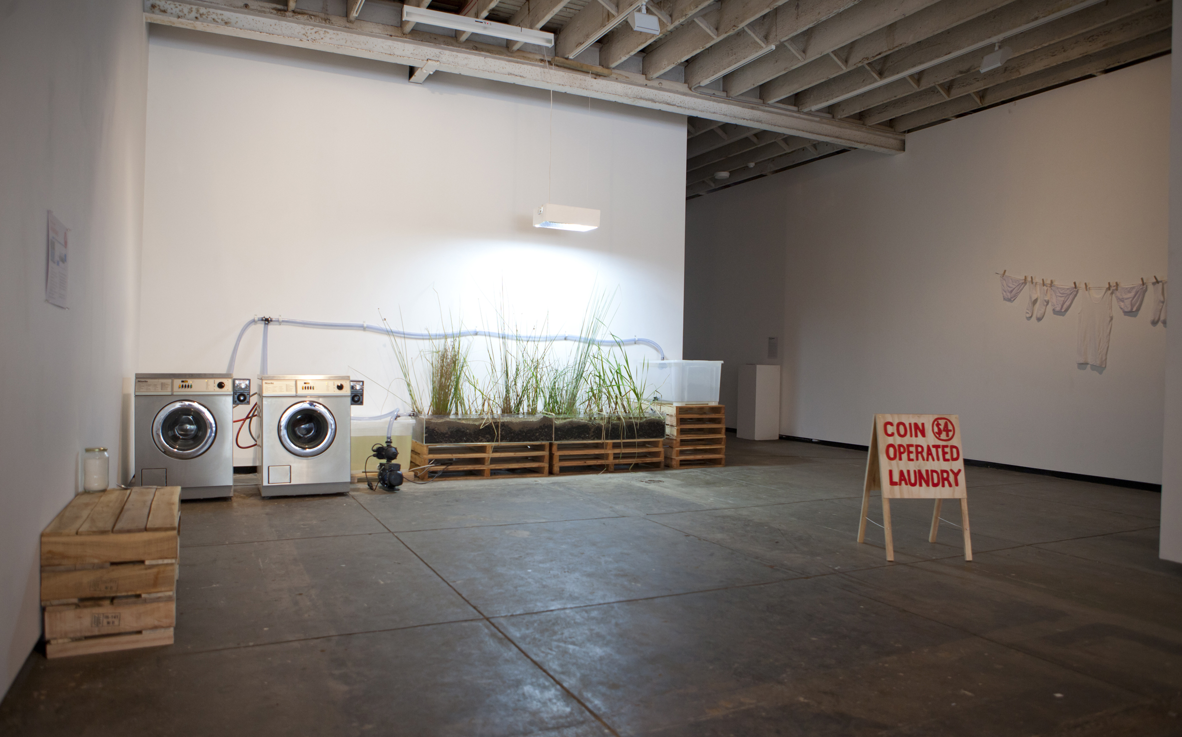Photo of an installation with two washers wired to a planter, with a sandwich board reading 'Coin operated laundry' in the foreground.