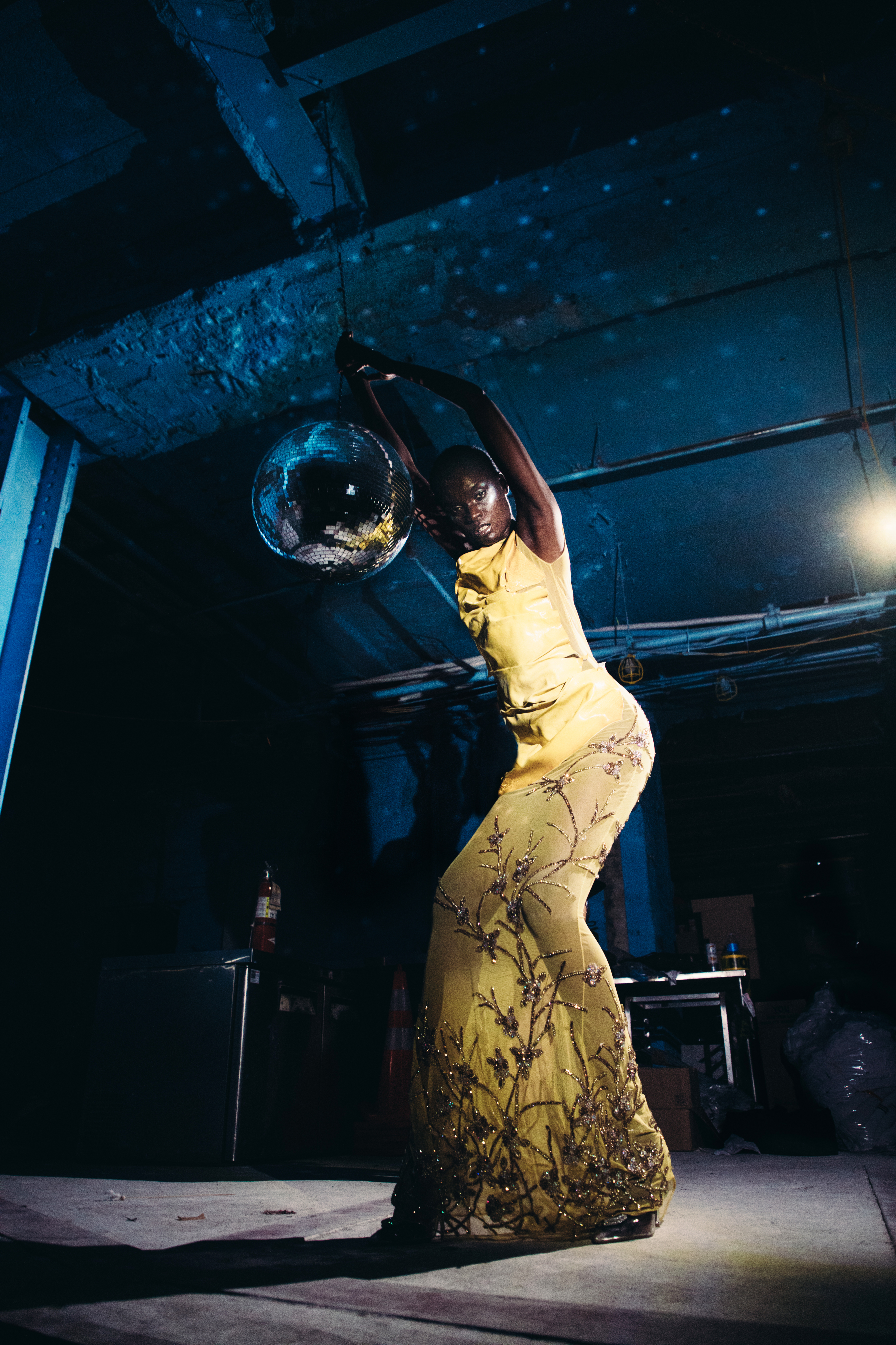  A Black model with their arms splayed out, one leg up, in a dancing posture.
     They are wearing a white draped headpiece with red and green drawings on the side,
     a shimmery dress with lavender pleating on the chest, diaphanous, flowy sheer pink
     fabric on the arms; these two fabrics flow together through the bottom half of the
     dress. The bodice is a beige color beaded with rhinestones, which shows through
     in the stomach area. The model is wearing earrings and has a septum piercing, and
     they wear black platform boots, against a white backdrop.