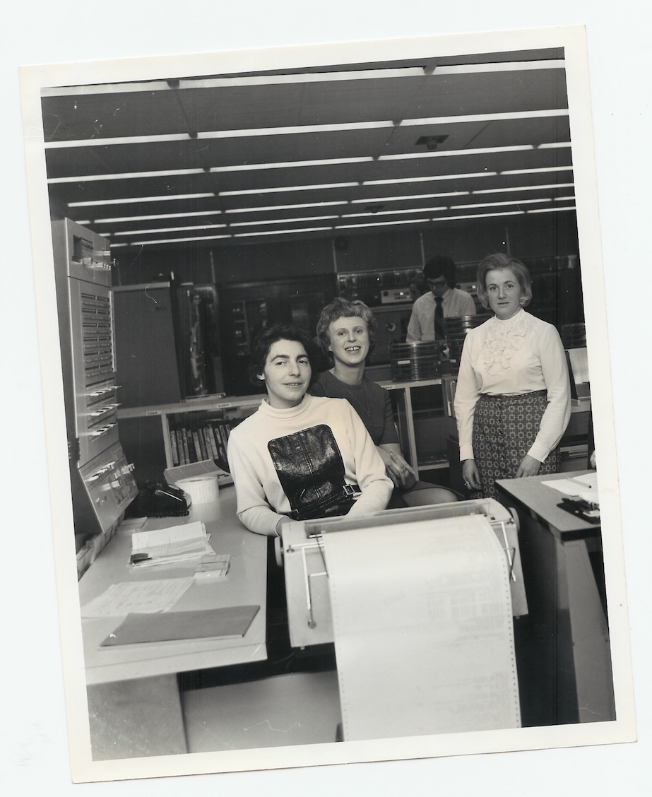 Steve Shirley, Ann Moffatt, and Dee Shermer pose among office computing equipment, with a large paper-fed machine in the foreground.
