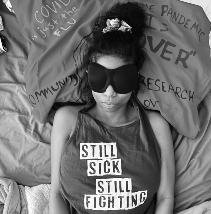 A person lies on bedding with an eye mask over their eyes. Their shirt reads ‘STILL SICK STILL FIGHTING.’ Handwritten words on the bedding include ‘COVID is just the FLU,’ ‘PANDEMIC,’ ‘COMMUNITY,’ and ‘RESEARCH.’